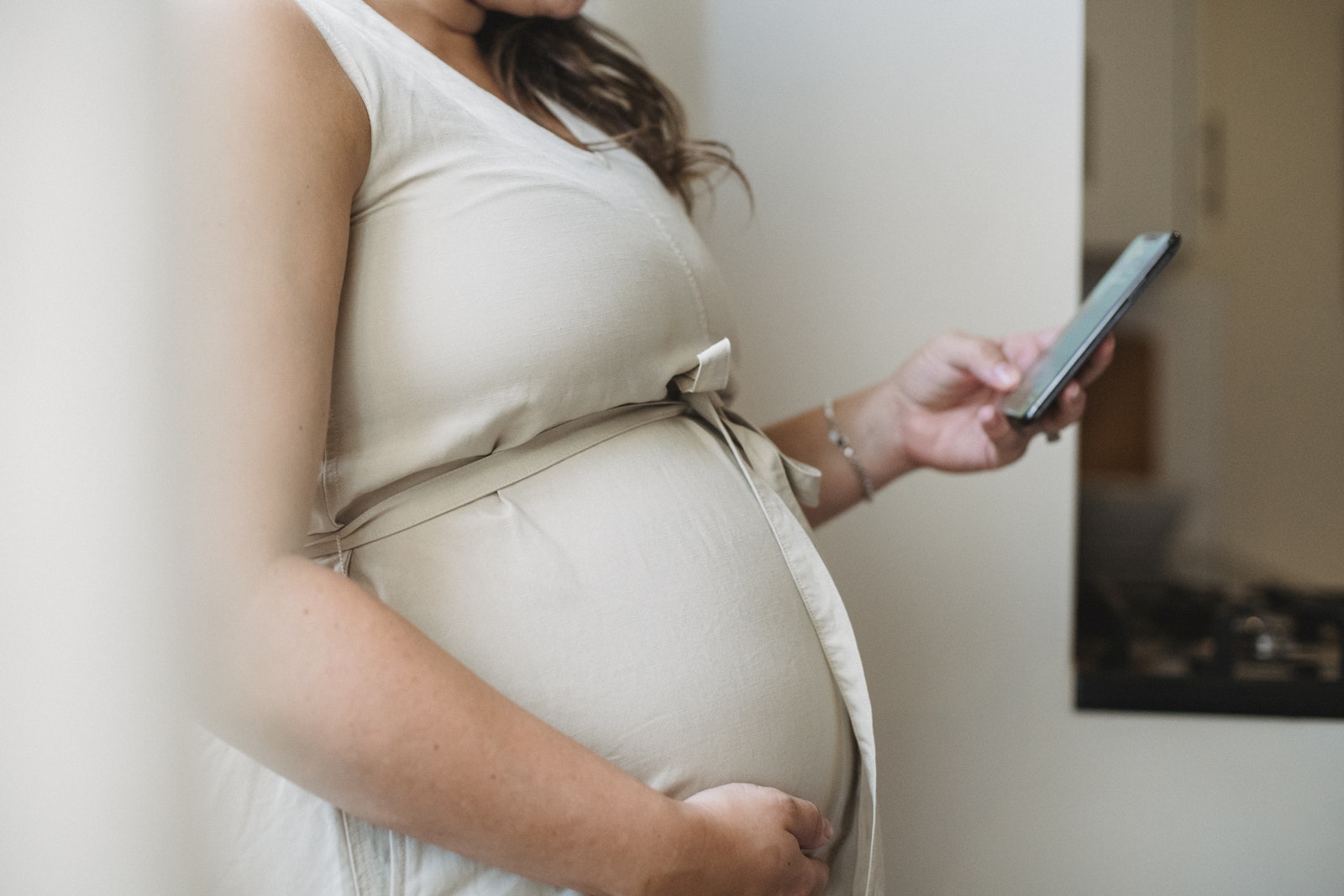 Side view crop unrecognizable pregnant woman text messaging on phone while standing in room in daylight