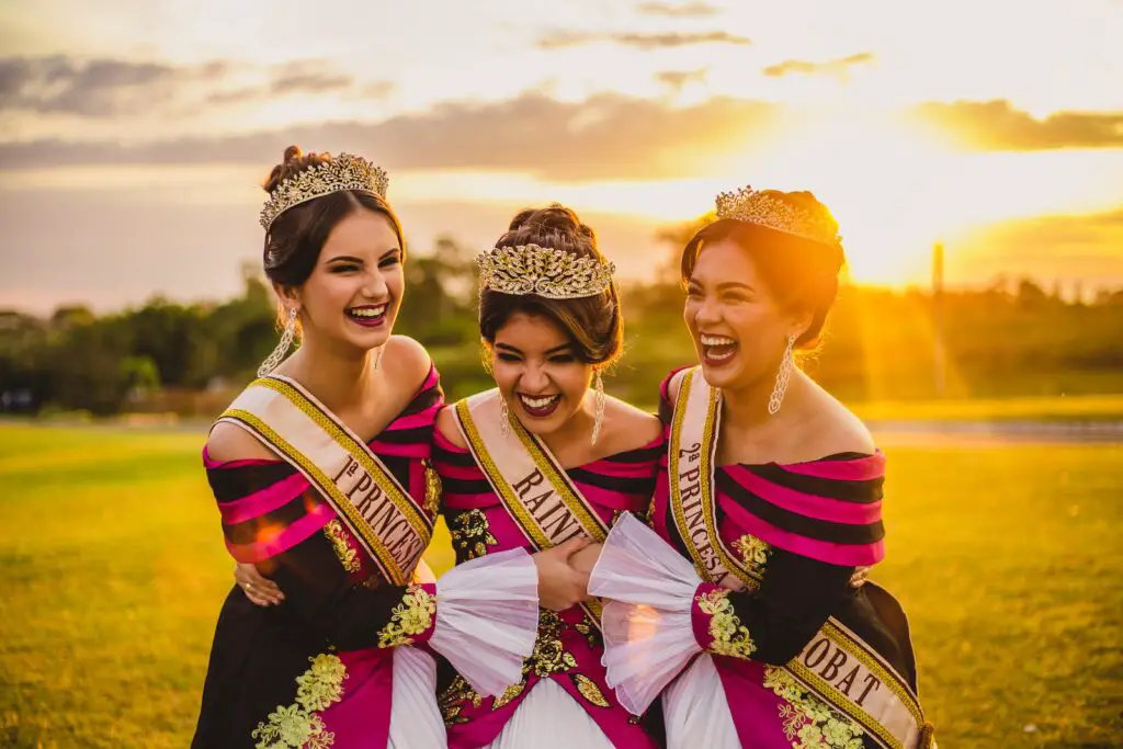 Laughing ethnic women in bright trendy apparel and crowns embracing on meadow under glowing cloudy sky at sunset in back lit