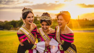 Laughing ethnic women in bright trendy apparel and crowns embracing on meadow under glowing cloudy sky at sunset in back lit