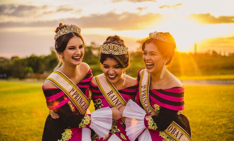 Laughing ethnic women in bright trendy apparel and crowns embracing on meadow under glowing cloudy sky at sunset in back lit