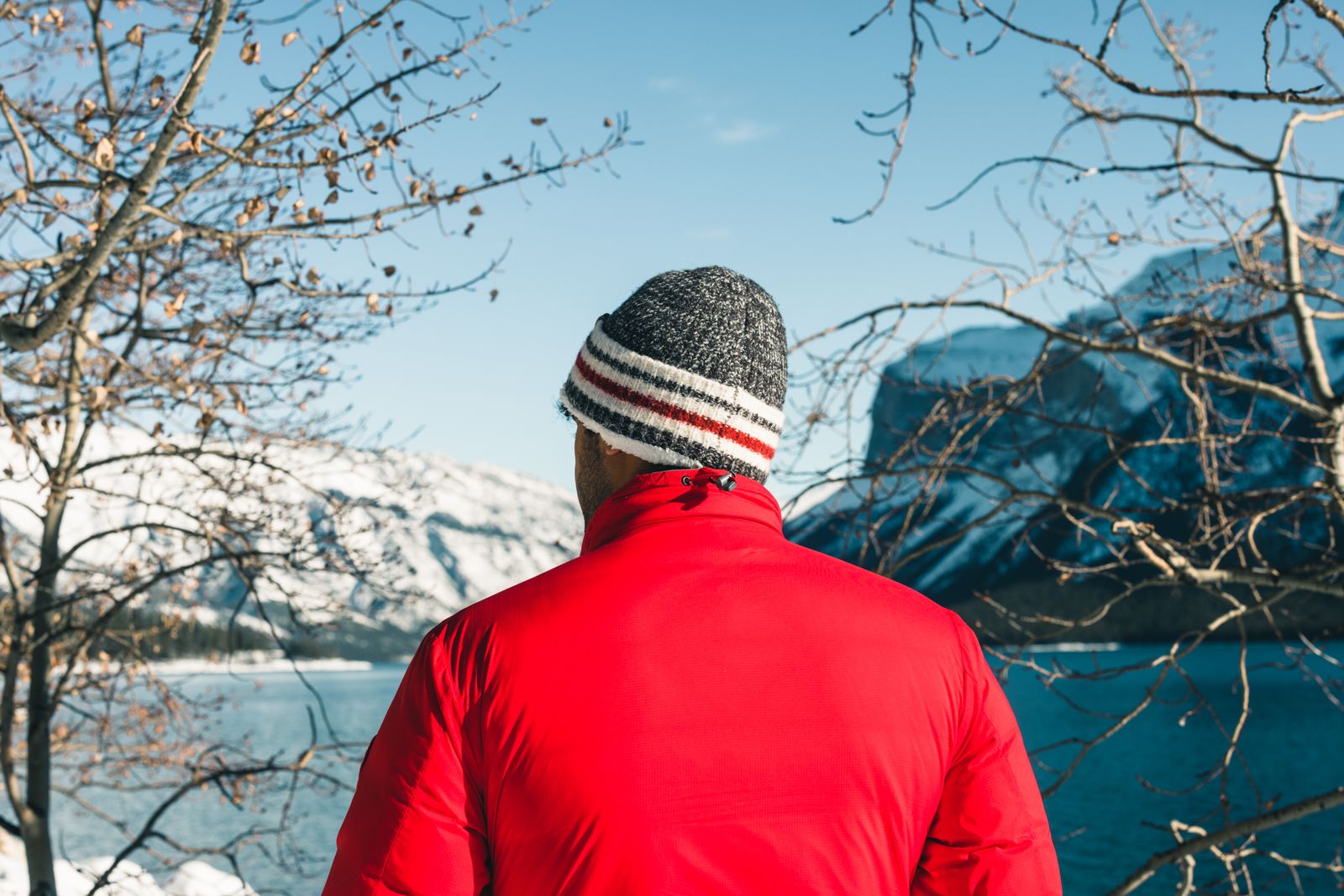 Uomo di spalle con berretto di lana e giacca rossa davanti a un lago di montagna innevato