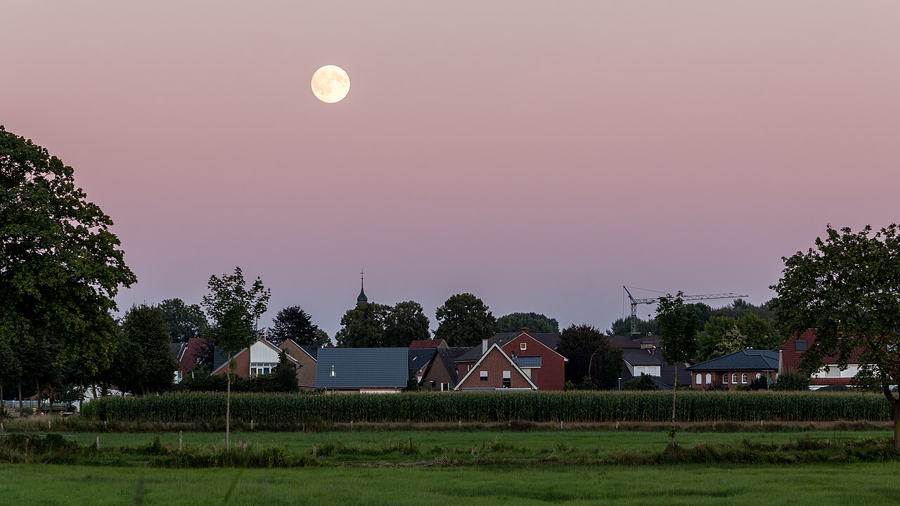 La Luna piena bassa nel cielo rosato del tramonto sopra i tetti e gli alberi di Hausdülmen, in Germania