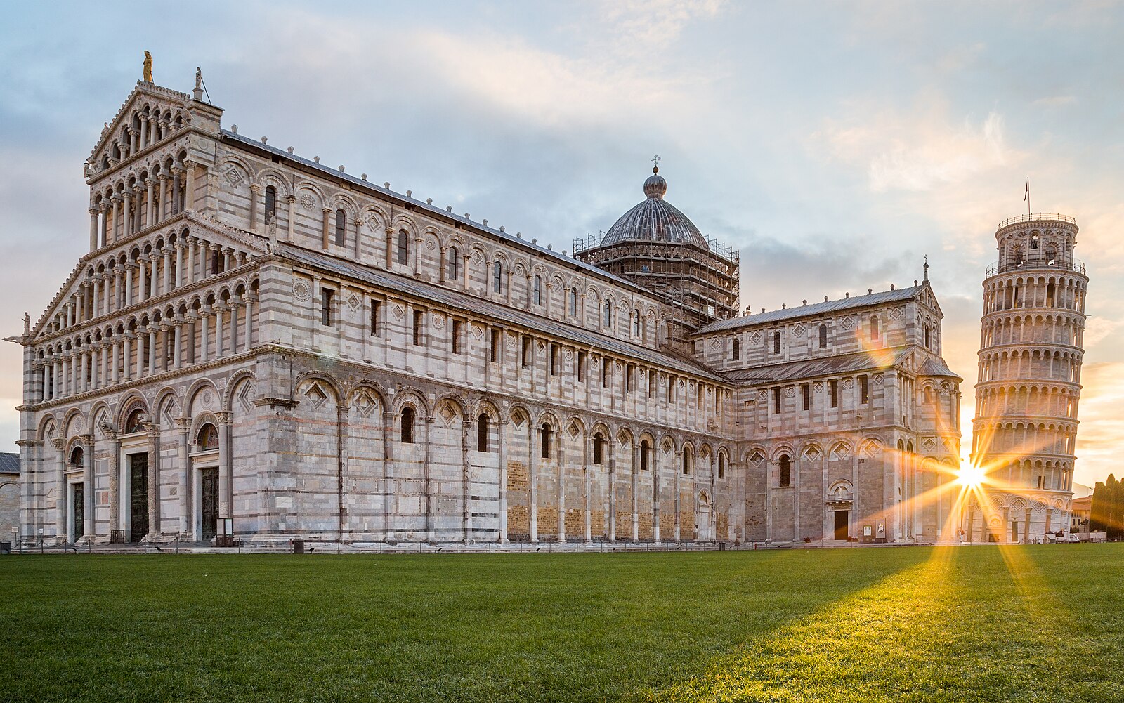 Il Duomo di Pisa e la Torre pendente all'alba in Piazza dei Miracoli, con il sole che spunta dietro la torre